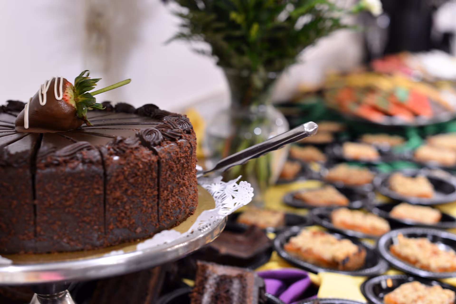 Close-up of a chocolate cake with a chocolate-covered strawberry on top, sliced and placed on a silver cake stand. In the background, there are multiple small plates with dessert servings arranged on a table with a yellow tablecloth and a vase with flowers.