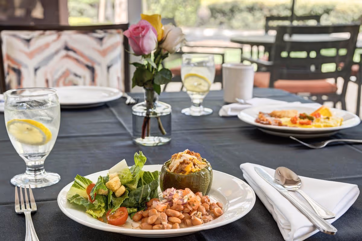 A dining table set with plates of food including a stuffed green bell pepper, salad with cherry tomatoes and croutons, and beans. There are two glasses of water with lemon slices, a white coffee cup, silverware wrapped in a white napkin, and a small vase with pink, yellow, and white roses. The background shows chairs and a window with a view of greenery outside.