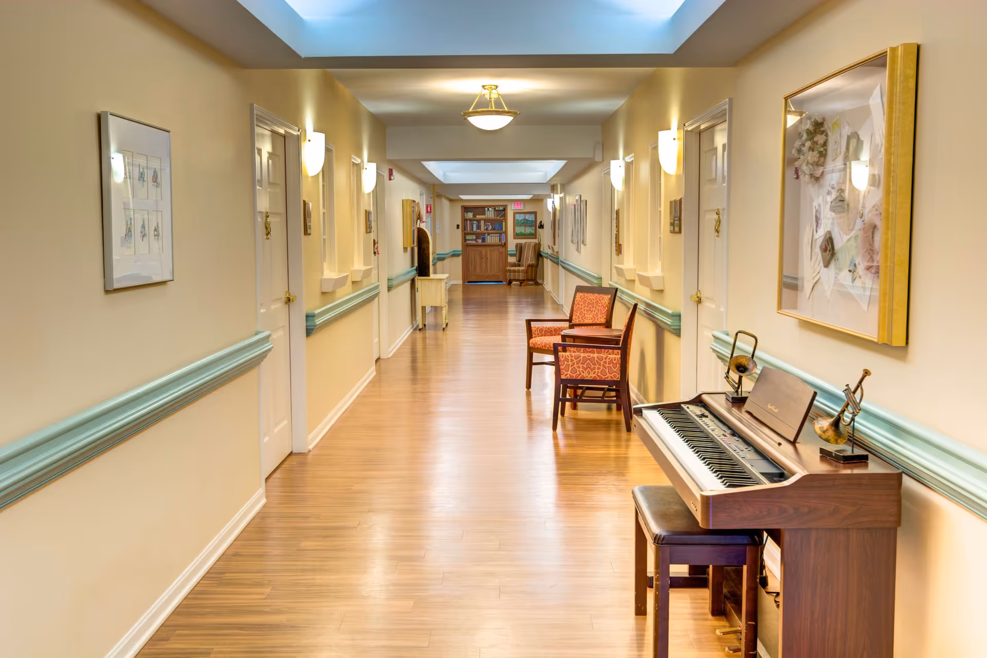 A well-lit hallway in a senior living facility with light yellow walls and wooden flooring. The hallway features several closed white doors on both sides, wall-mounted lights, framed artwork, and a wooden electronic piano with a bench on the right side. Two patterned armchairs are placed further down the hallway near a small table, and a bookshelf is visible at the end of the corridor.