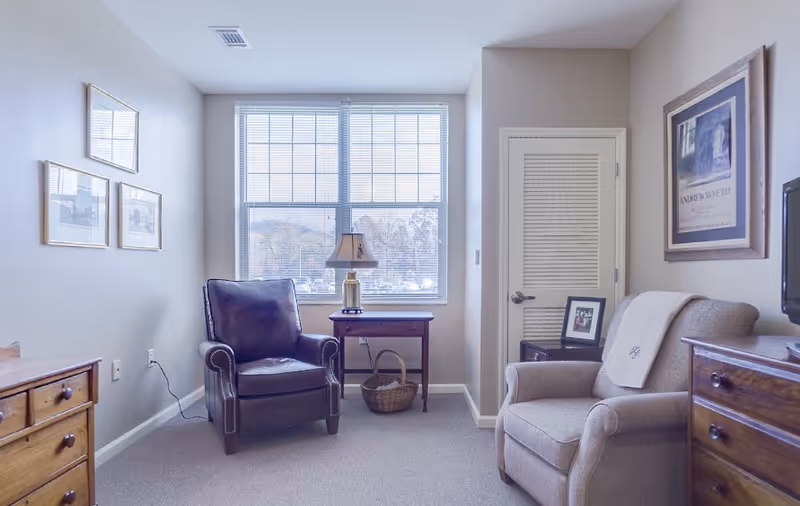 A cozy living room area with two armchairs, one dark leather and one beige fabric, positioned near a window with blinds. A small wooden table with a lamp and a basket underneath sits between the chairs. The walls are decorated with framed pictures and a door with a louvered panel is visible.
