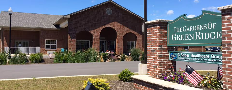 Front exterior of a brick senior living building with a sign reading "The Gardens of Green Ridge", landscaped beds, and American flags.