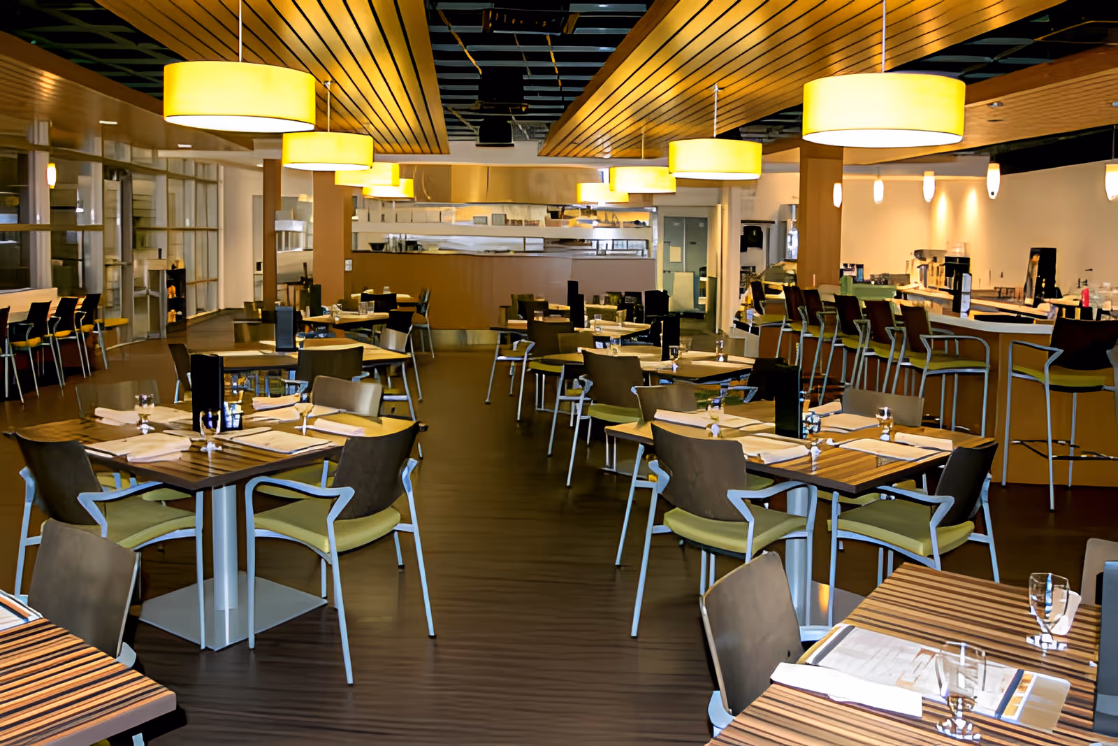 A modern dining room with multiple wooden tables and chairs arranged neatly. The tables are set with placemats, napkins, glasses, and menus. There are large round pendant lights hanging from a wooden slatted ceiling. A bar area with high stools is visible on the right side, and the back of the room features a kitchen or serving area with stainless steel equipment.