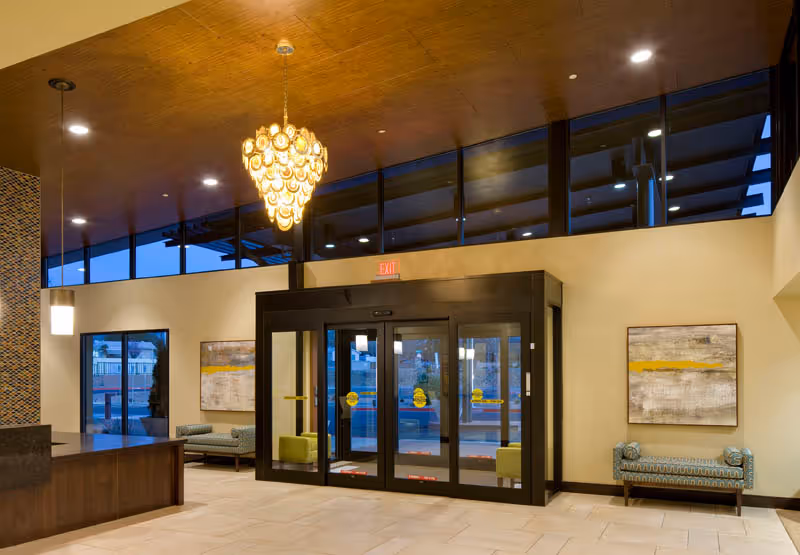 Interior view of a senior living facility lobby with a modern chandelier hanging from a wooden ceiling. The lobby features large glass automatic sliding doors, two patterned benches on either side, abstract artwork on the walls, and a reception desk on the left side.
