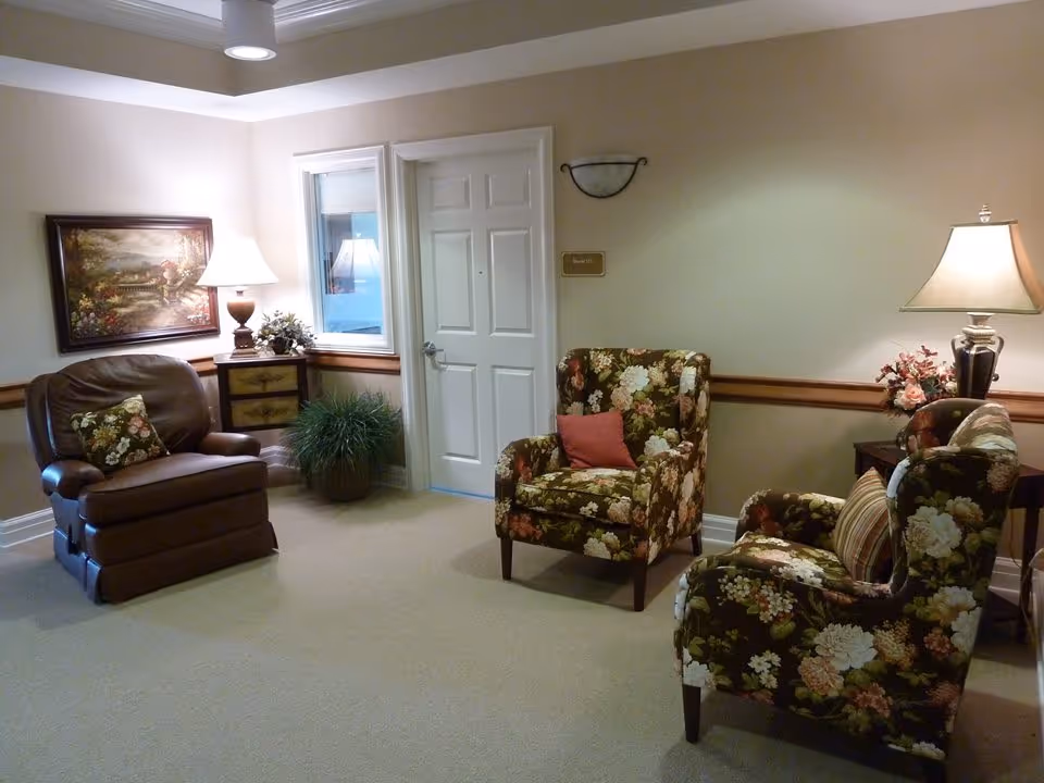 Small lounge with floral upholstered chairs, a leather recliner, side tables with lamps, a potted plant, and a closed white door.