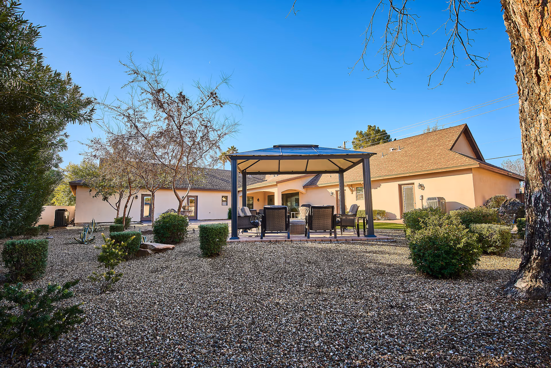 Outdoor garden area of a senior living facility with a gazebo featuring several chairs and a table underneath. The surrounding yard has gravel ground cover, trimmed bushes, and leafless trees. The building in the background has a beige exterior and a brown roof under a clear blue sky.
