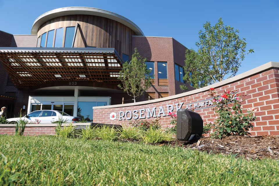 Exterior view of the Rosemark At Mayfair Park building with a brick facade, large windows, and a curved wooden architectural feature above the entrance. In front, there is a brick sign with the facility name surrounded by landscaping with green grass, plants, and flowers under a clear blue sky.