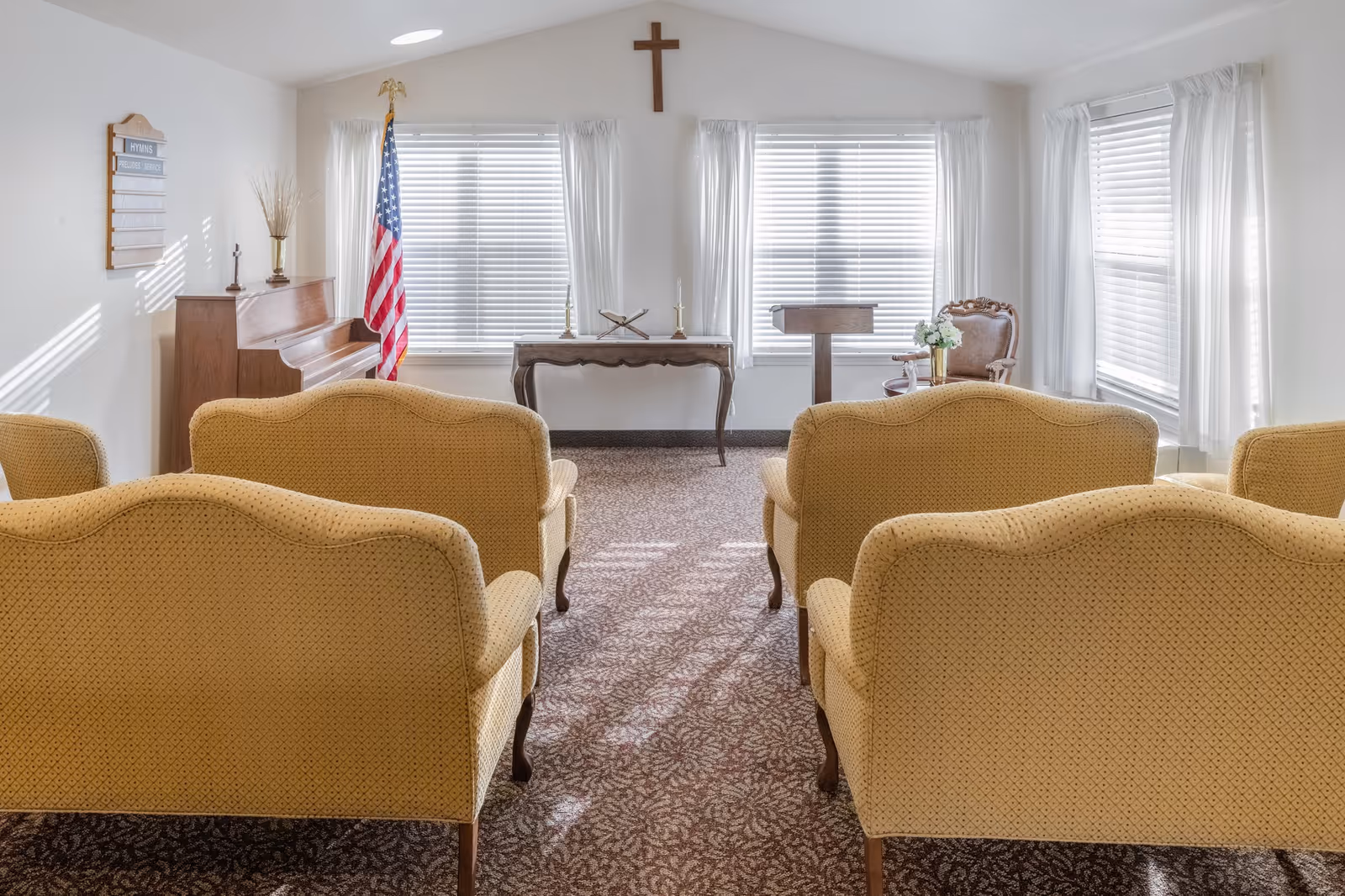 A small chapel or meeting room with rows of yellow upholstered chairs facing a wooden piano, an American flag, a wooden table with candles and a Bible, a lectern, and a cross mounted on the wall above three windows with white blinds and curtains.