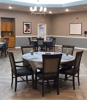 Communal dining room with round tables set with napkins and place settings, chairs, a chandelier, and wall artwork.