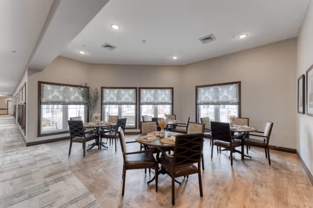 A bright dining area in a senior living facility with round wooden tables set for meals and cushioned chairs. Large windows with patterned blinds allow natural light to fill the room. The floor is a combination of wood and carpet, and the walls are painted in a neutral tone.