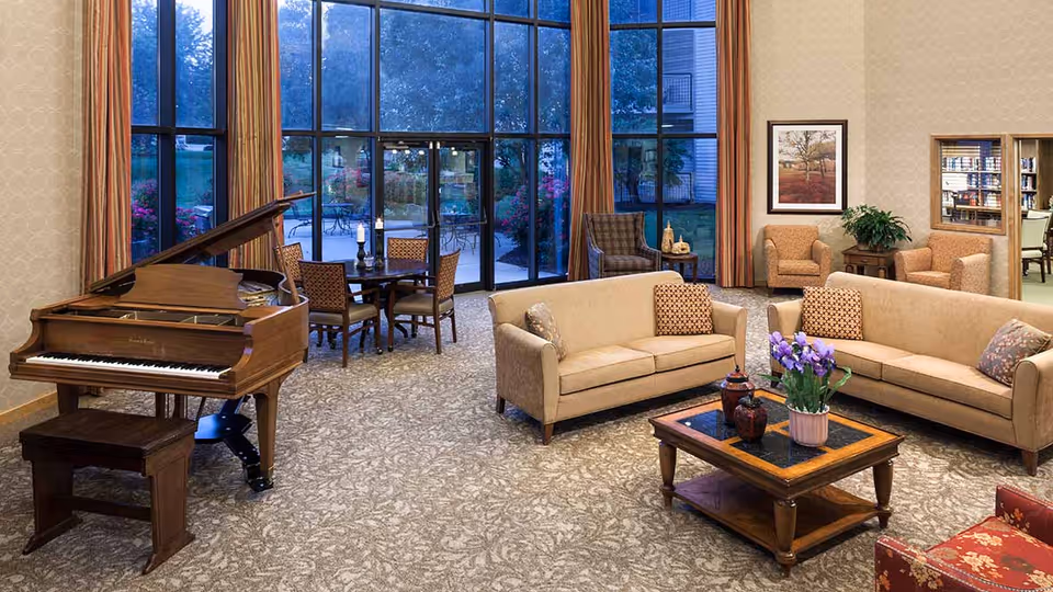 A spacious living room area with large floor-to-ceiling windows showing an outdoor patio and garden. The room features a grand piano with a bench, two beige sofas with patterned cushions, a wooden coffee table with a flower pot and decorative items, a small round dining table with four chairs, and additional armchairs near the walls. The walls have framed artwork and the room is carpeted with a patterned design.