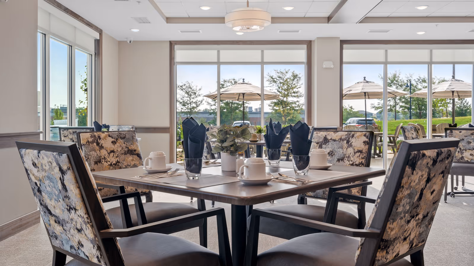 A bright dining area with a square wooden table set for four, featuring patterned cushioned chairs, white cups and saucers, glasses with black napkins, and a small potted plant centerpiece. Large windows in the background reveal an outdoor patio with umbrellas and greenery.