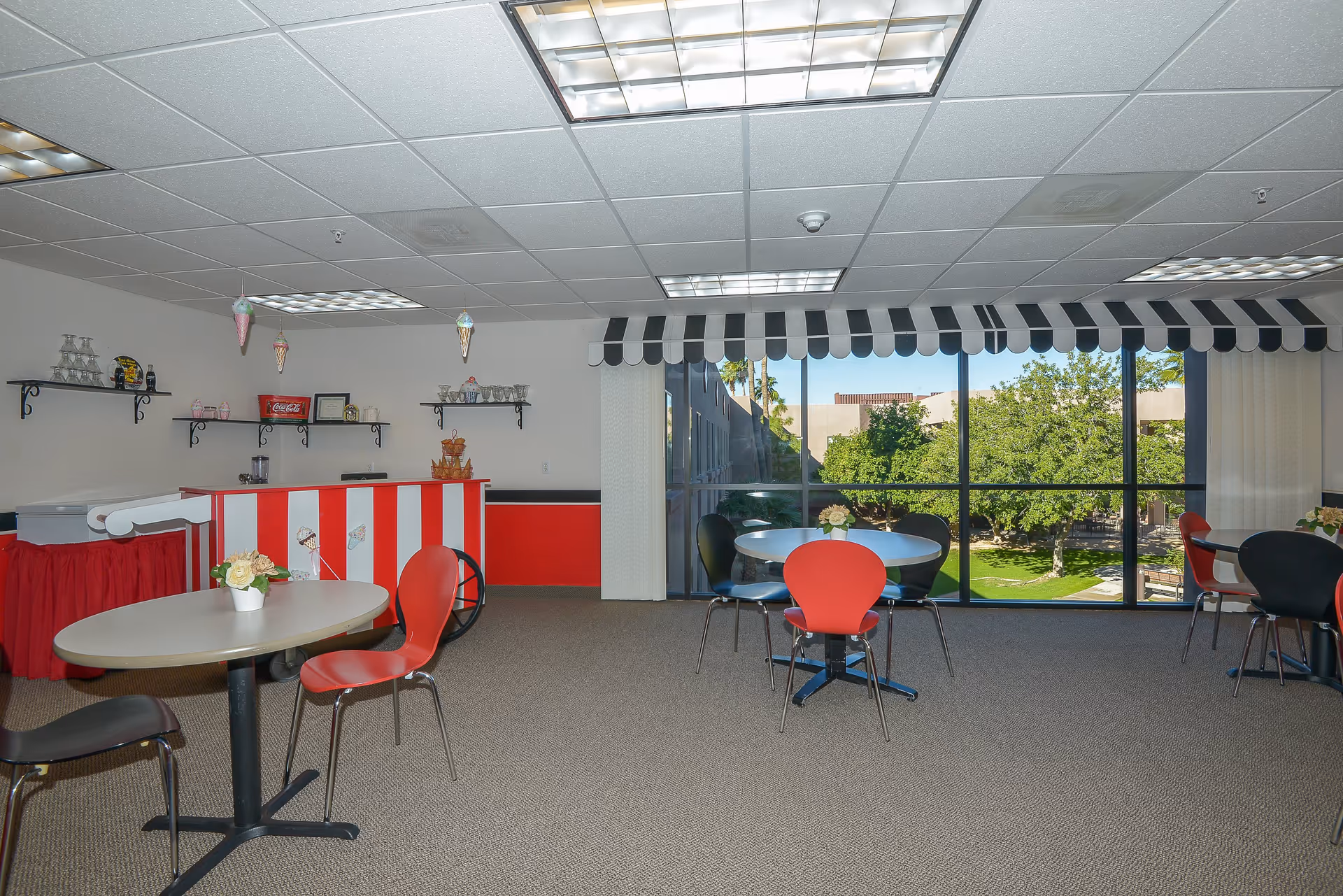 Interior view of a common dining area with several tables and chairs. The room features a red and white striped counter decorated with ice cream cone hanging ornaments. Large windows with black and white striped valances provide a view of green trees and a courtyard outside.