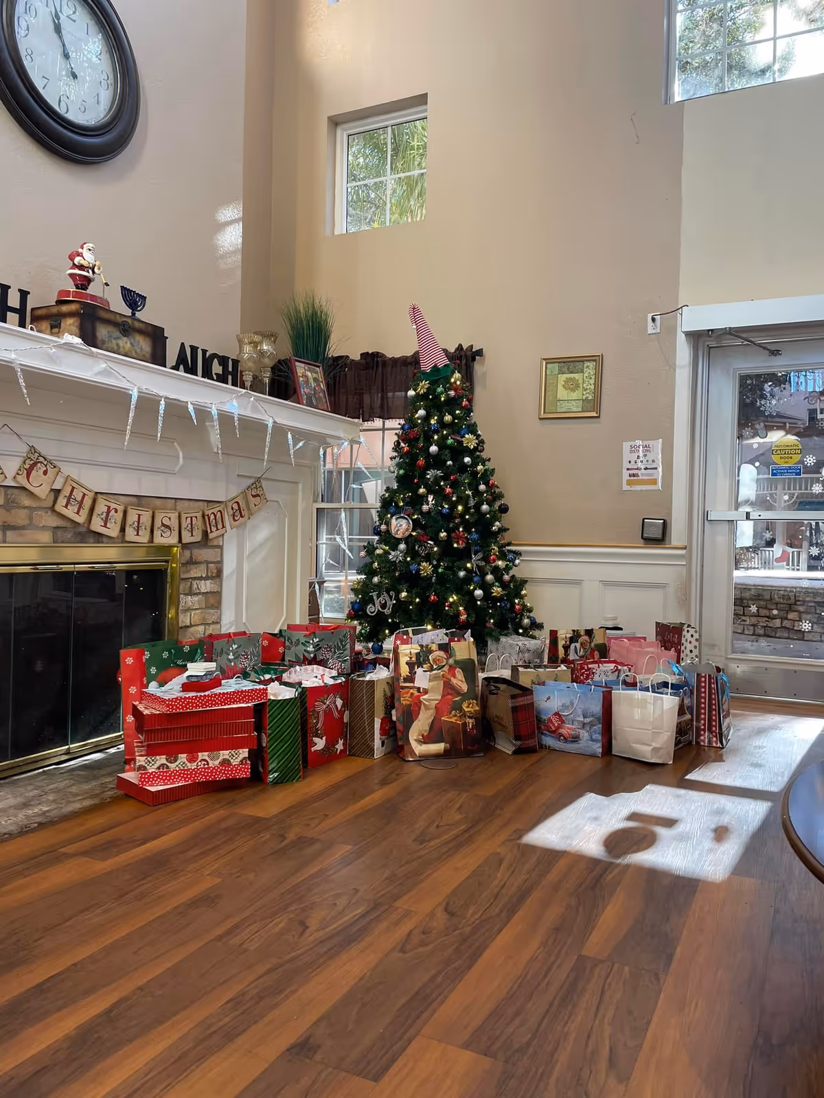 Interior of a living room decorated for Christmas with a decorated Christmas tree and numerous wrapped gifts placed on the wooden floor. A fireplace with a mantel decorated with a Santa figurine, a menorah, and a Christmas banner is visible. There is a large clock on the wall and windows letting in natural light.