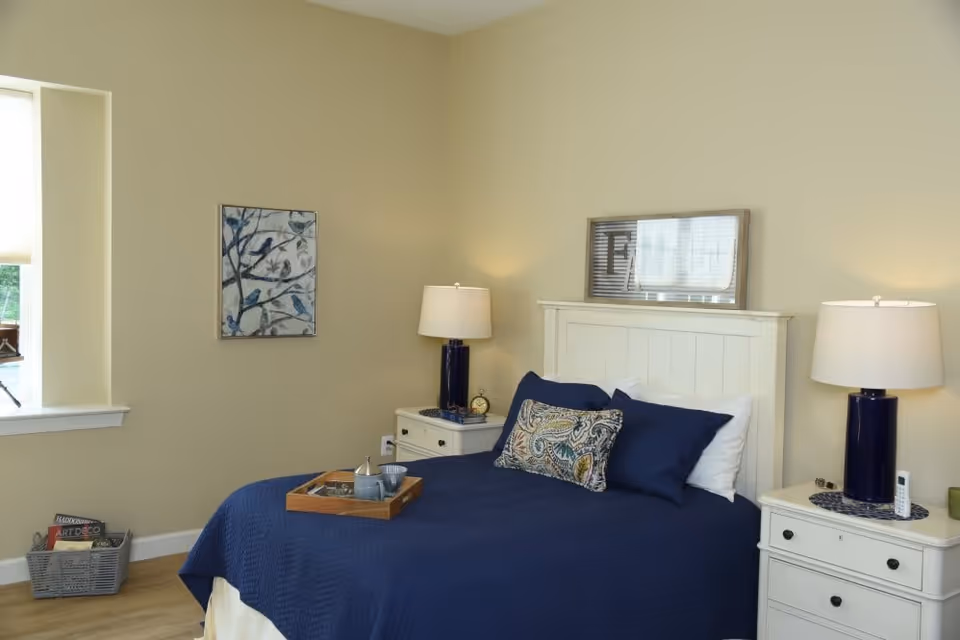 A cozy bedroom with a white wooden bed frame and a navy blue bedspread. There are two white nightstands on either side of the bed, each with a lamp featuring a dark blue base and a white lampshade. A decorative pillow with a paisley pattern is placed on the bed along with two white pillows. A wooden tray with a teapot and cup is on the bed. The walls are painted light beige, and there is a framed artwork of birds on a branch hanging on the wall. A window with a white blind is on the left side, and a basket with books is on the floor near the window.