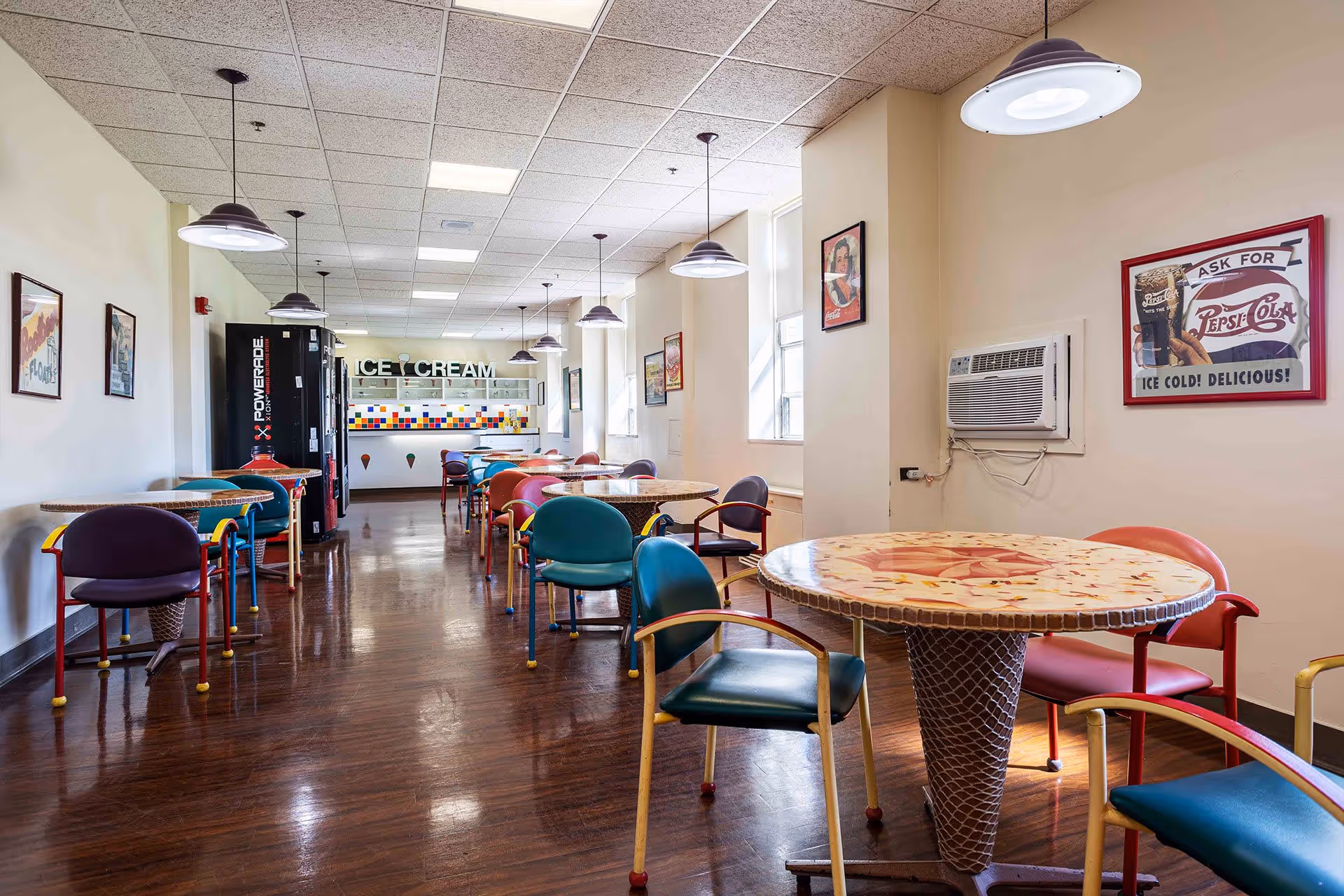 A brightly lit dining area with multiple round tables and colorful chairs arranged neatly. The floor is wooden, and the ceiling has several hanging light fixtures. At the far end, there is a counter with a sign that reads 'ICE CREAM' above it, and a vending machine is visible on the left side. The walls are decorated with vintage-style posters, and a window air conditioning unit is mounted on the right wall.