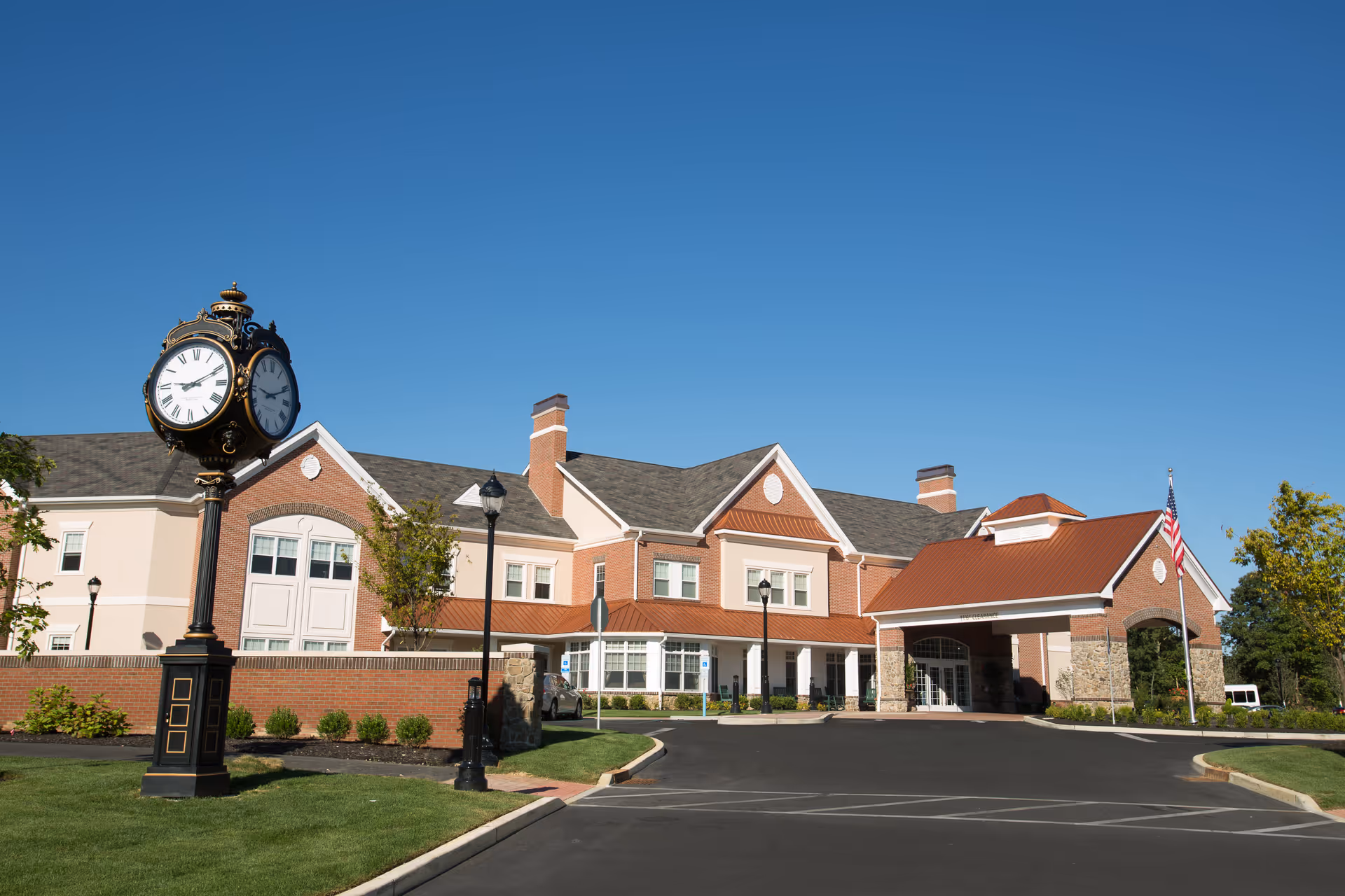 Front exterior of a brick senior living building with a decorative street clock, covered entrance, and American flag under a clear blue sky.