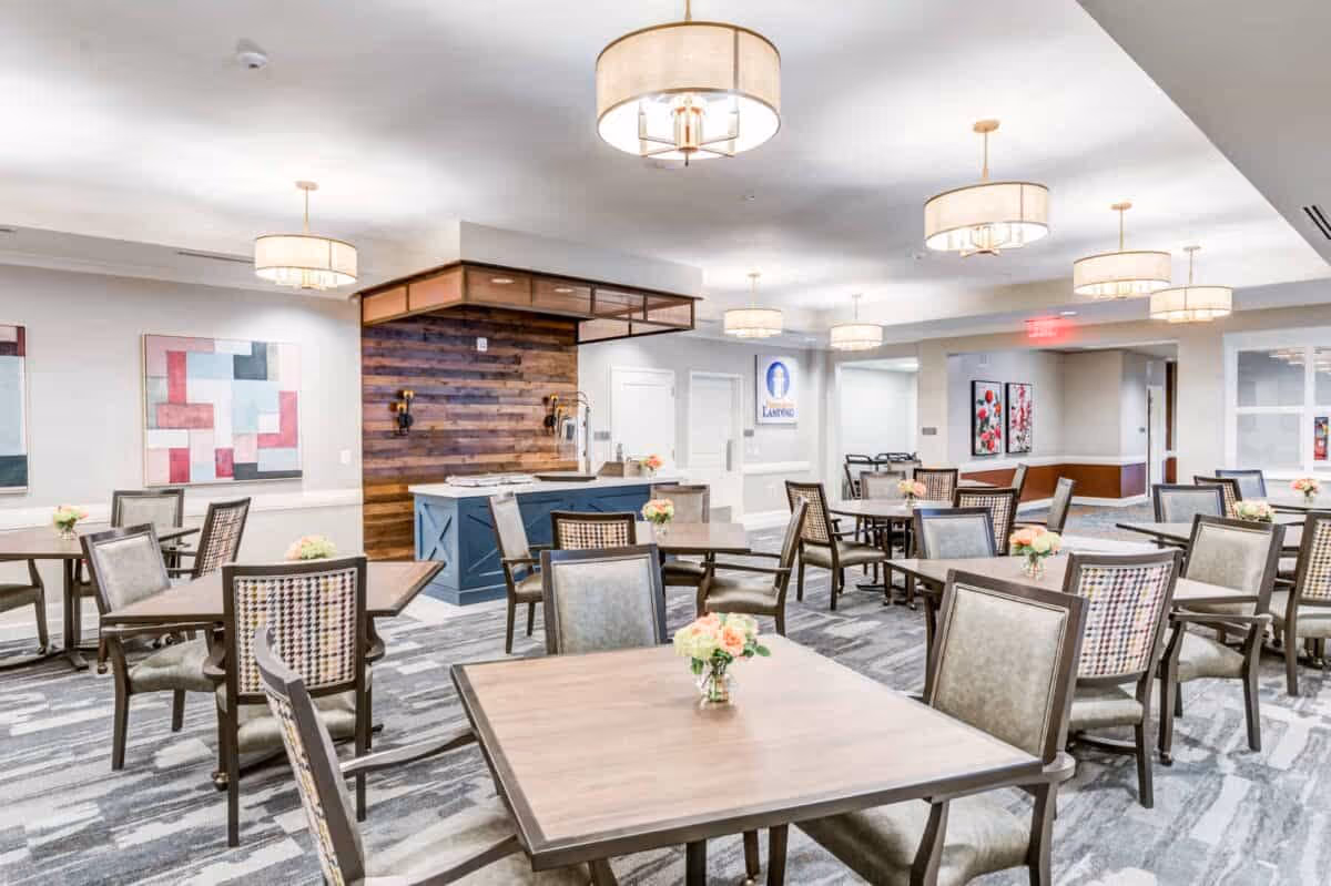 A bright and spacious dining room with multiple square wooden tables and cushioned chairs arranged neatly. Each table has a small vase with flowers. The room features modern pendant lights hanging from the ceiling, abstract artwork on the walls, and a wooden accent wall with a counter area in the background.