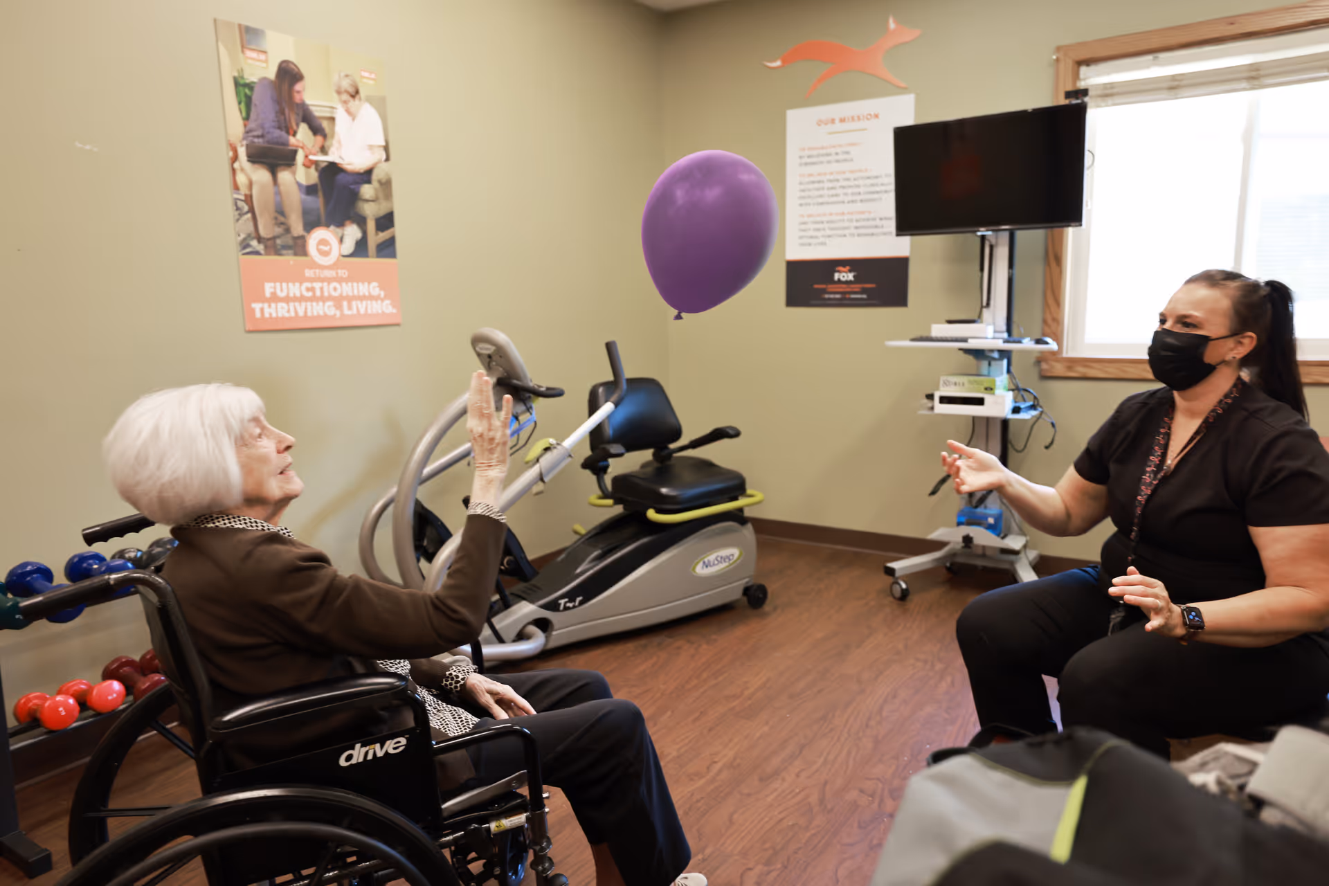 An elderly woman in a wheelchair playing with a purple balloon with a caregiver wearing a black mask in a room with exercise equipment and motivational posters on the walls.