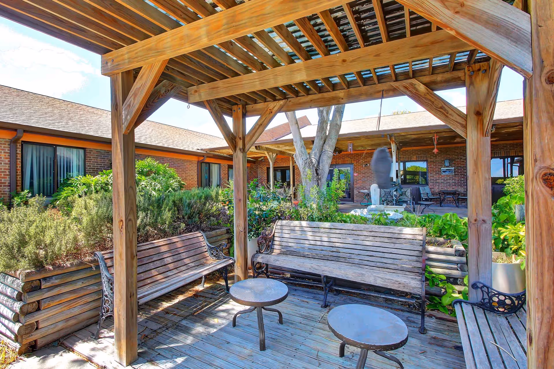 Outdoor seating area with wooden benches and small round tables under a wooden pergola. Surrounding the area are green plants and shrubs, with a brick building in the background featuring windows and doors.