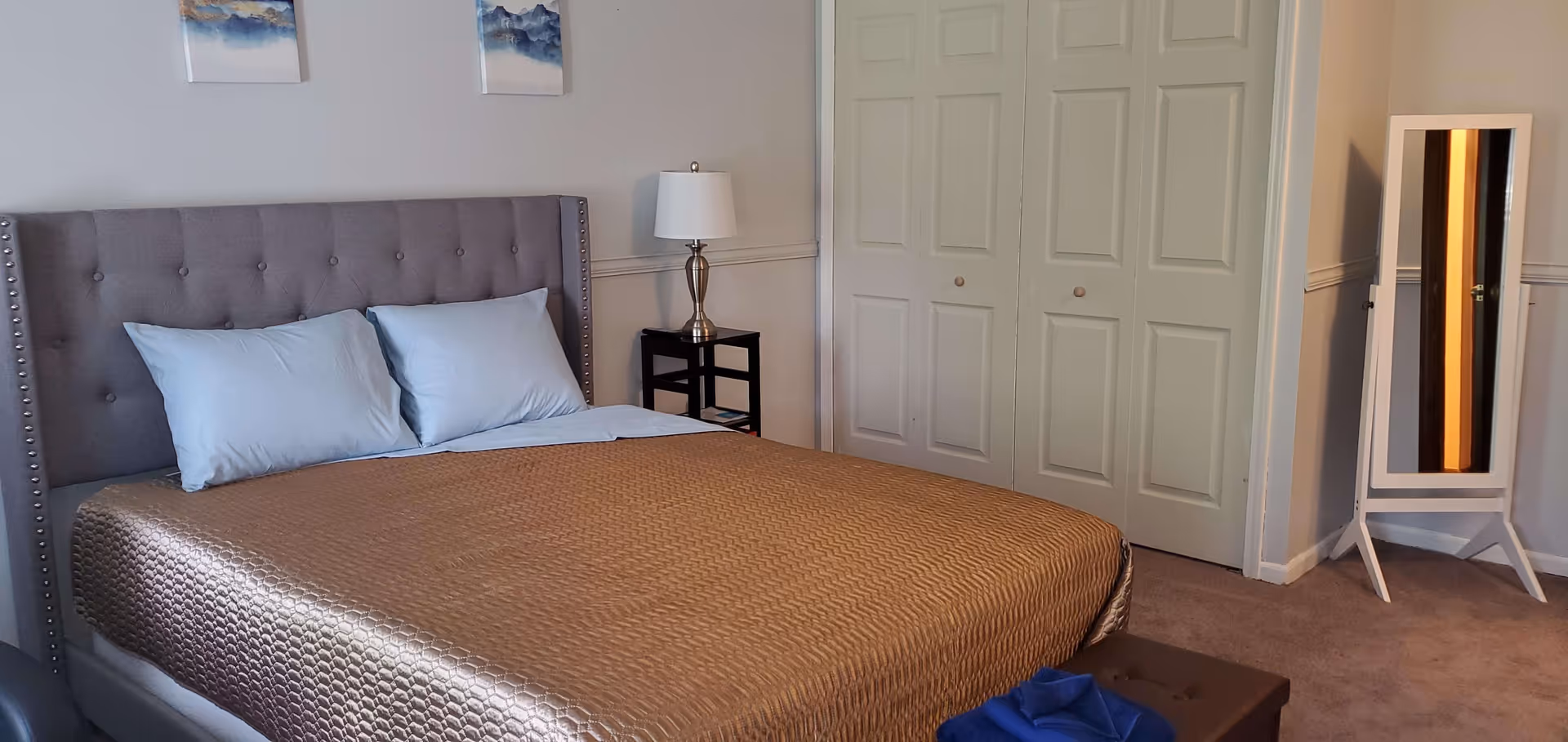 A bedroom with a gray upholstered bed with two light blue pillows and a gold textured bedspread. There is a small black nightstand with a silver lamp on it next to the bed. Two abstract paintings hang on the wall above the bed. A white standing mirror is positioned near a closed white closet with paneled doors. The room has beige carpet and light-colored walls.