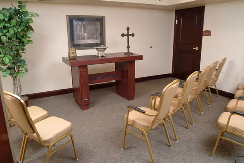 A small chapel or meditation room with beige cushioned chairs arranged in rows facing a wooden altar table. The altar holds a cross, a framed picture, and a decorative lantern. There is a potted plant in the corner and a closed wooden door on the right side.