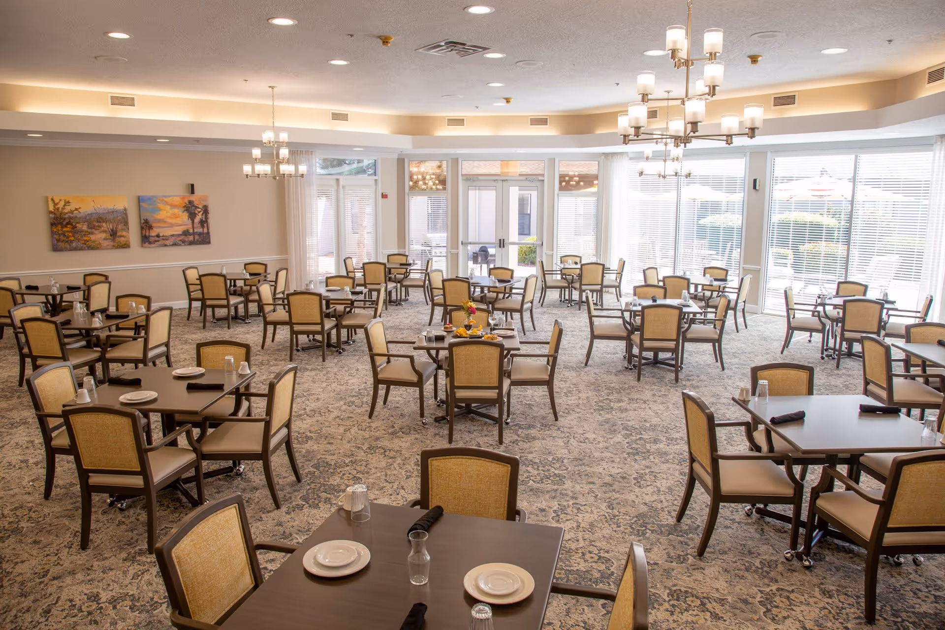 A spacious dining room with multiple dark wood tables and beige cushioned chairs arranged neatly. Each table is set with plates, glasses, and black napkins. Large windows with blinds allow natural light to fill the room. Two chandeliers hang from the ceiling, and there are two landscape paintings on the far wall. The room has a patterned carpet and a bright, welcoming atmosphere.