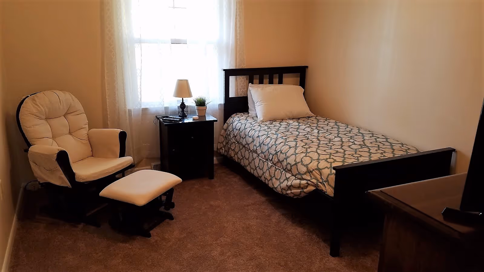 A small bedroom with a single bed covered in a patterned comforter and a white pillow. Next to the bed is a dark wooden nightstand with a lamp, a small plant, and a remote control. To the left of the nightstand is a cushioned armchair with a matching footrest. A window with sheer white curtains is behind the nightstand and chair. The room has beige walls and carpeted floor.