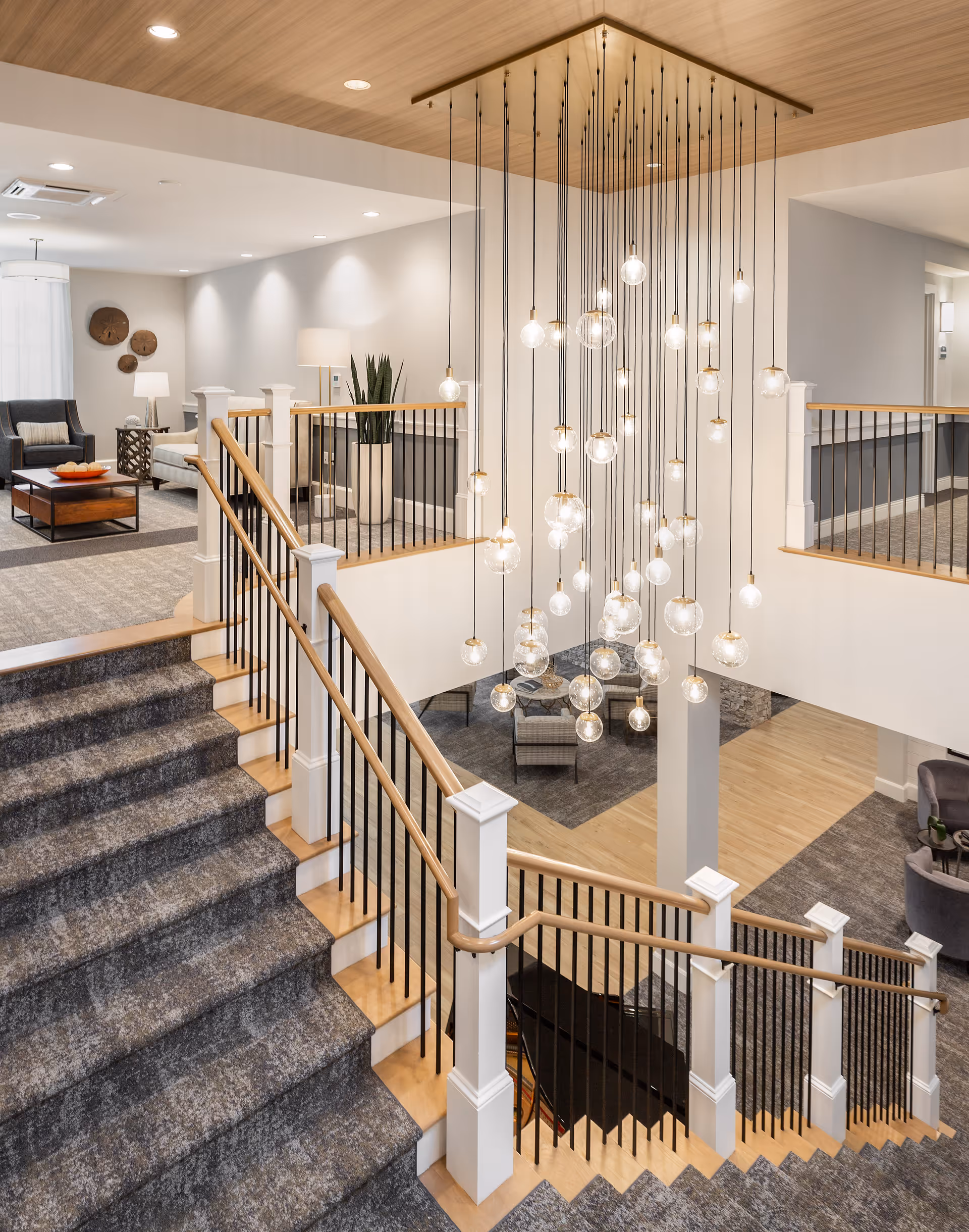 Interior view of a senior living facility featuring a carpeted staircase with wooden handrails and black balusters. A modern chandelier with multiple hanging light bulbs is suspended from the ceiling above the stairwell. The surrounding area includes seating arrangements with chairs, a sofa, side tables, lamps, and decorative wall art. The space is well-lit with recessed ceiling lights and has a warm, inviting atmosphere.