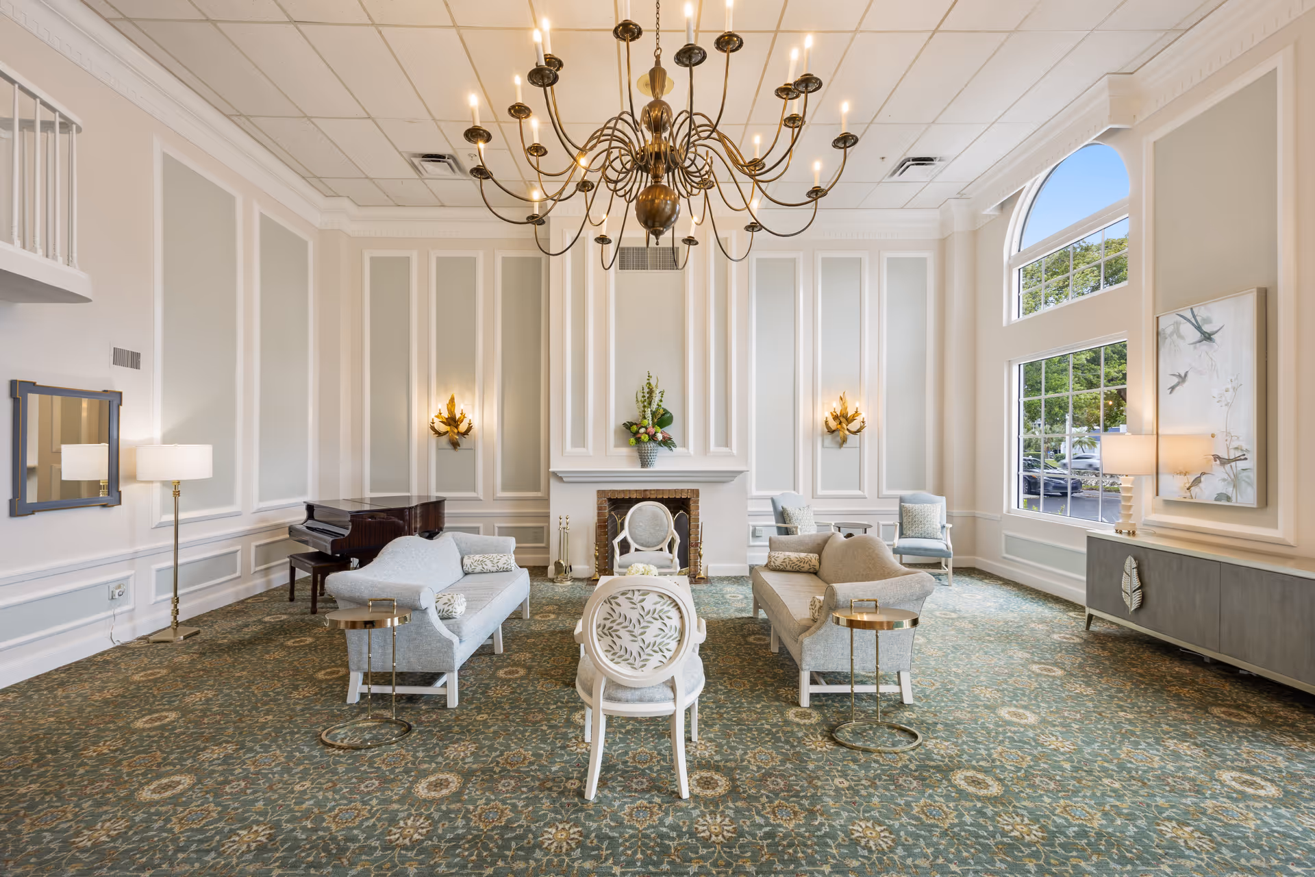 A spacious and elegant senior living facility lounge with high ceilings, a large chandelier, patterned carpet, two light gray sofas facing each other, a white chair with floral upholstery, a fireplace with floral arrangement on the mantel, a grand piano in the corner, large windows letting in natural light, and soft wall sconces.