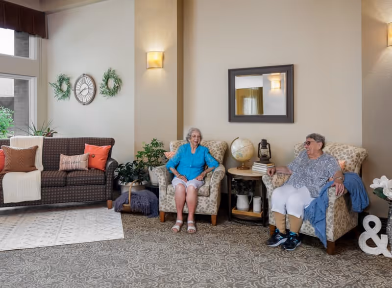 Two elderly women sitting and smiling in a well-lit living room area with patterned armchairs, a side table with a globe and books, a large mirror on the wall, and a sofa with decorative pillows and a throw blanket. The room has carpeted flooring and wall sconces providing warm lighting.