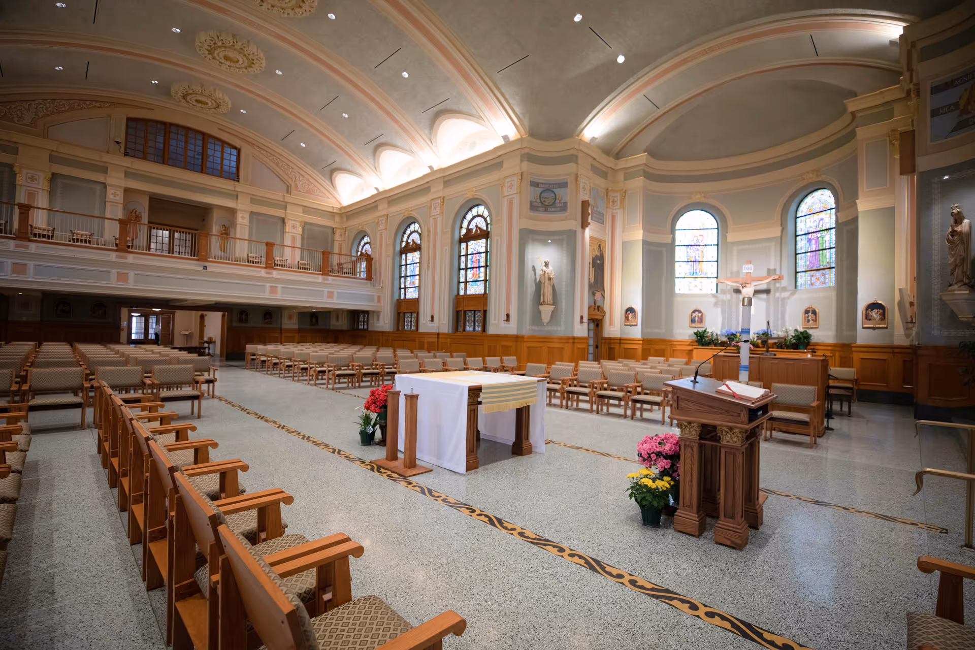 Interior view of a chapel with rows of wooden chairs arranged facing an altar covered with a white cloth. The chapel features high arched ceilings with decorative molding, stained glass windows, religious statues, and a lectern with a microphone and an open book. There are potted flowers near the altar and lectern.