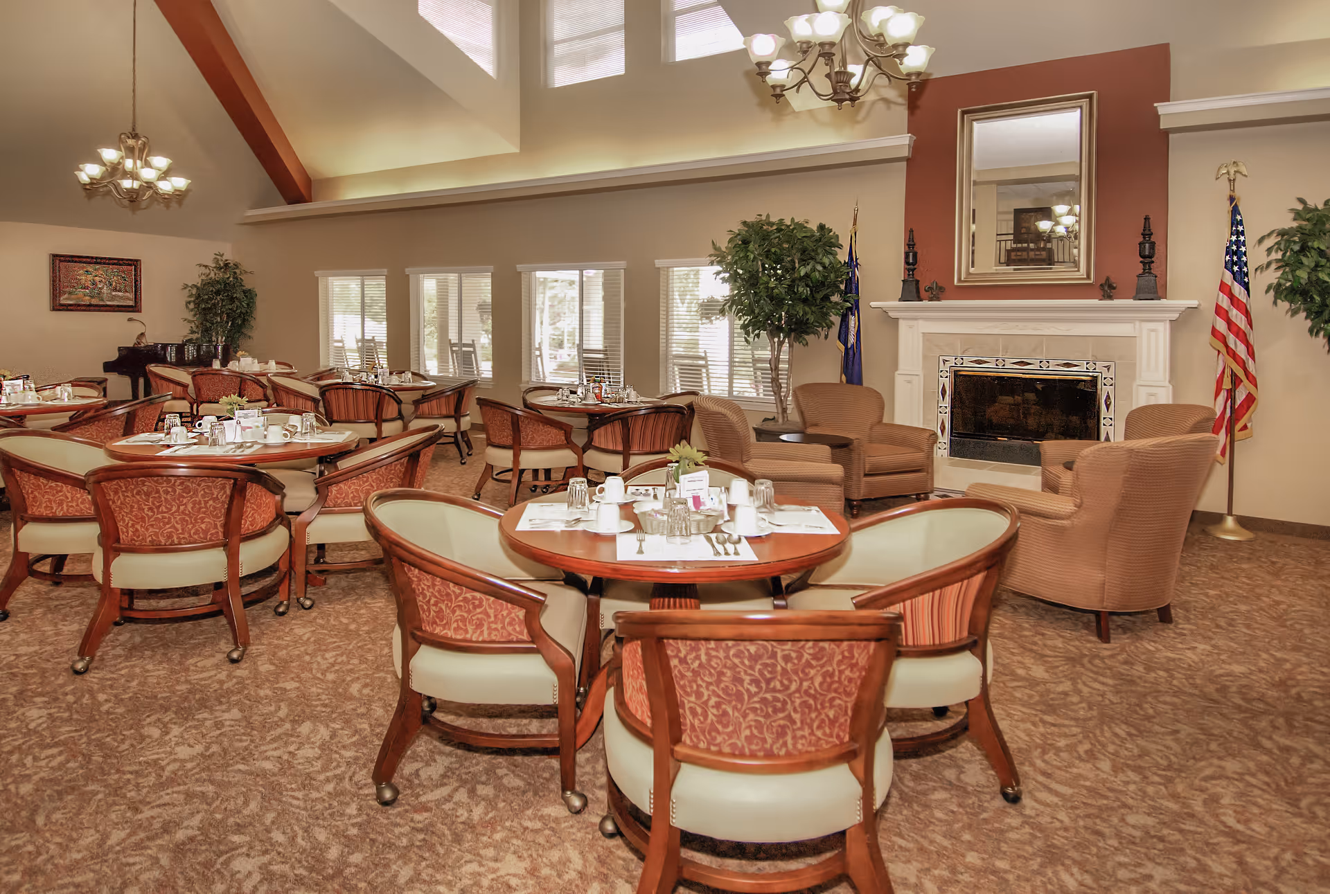 A spacious dining room with multiple round tables set with cups, glasses, and utensils. The room features upholstered chairs with wooden frames, a fireplace with a large mirror above it, two flags on either side, and several potted plants. Large windows allow natural light to fill the room, and chandeliers hang from the ceiling.
