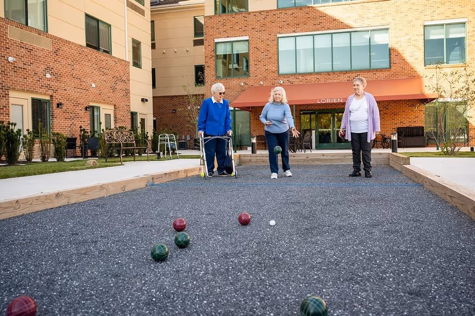 Three elderly women playing bocce ball on an outdoor court in front of a brick building with a red awning labeled 'Lorien'. One woman uses a walker, and the other two are standing and watching the game.