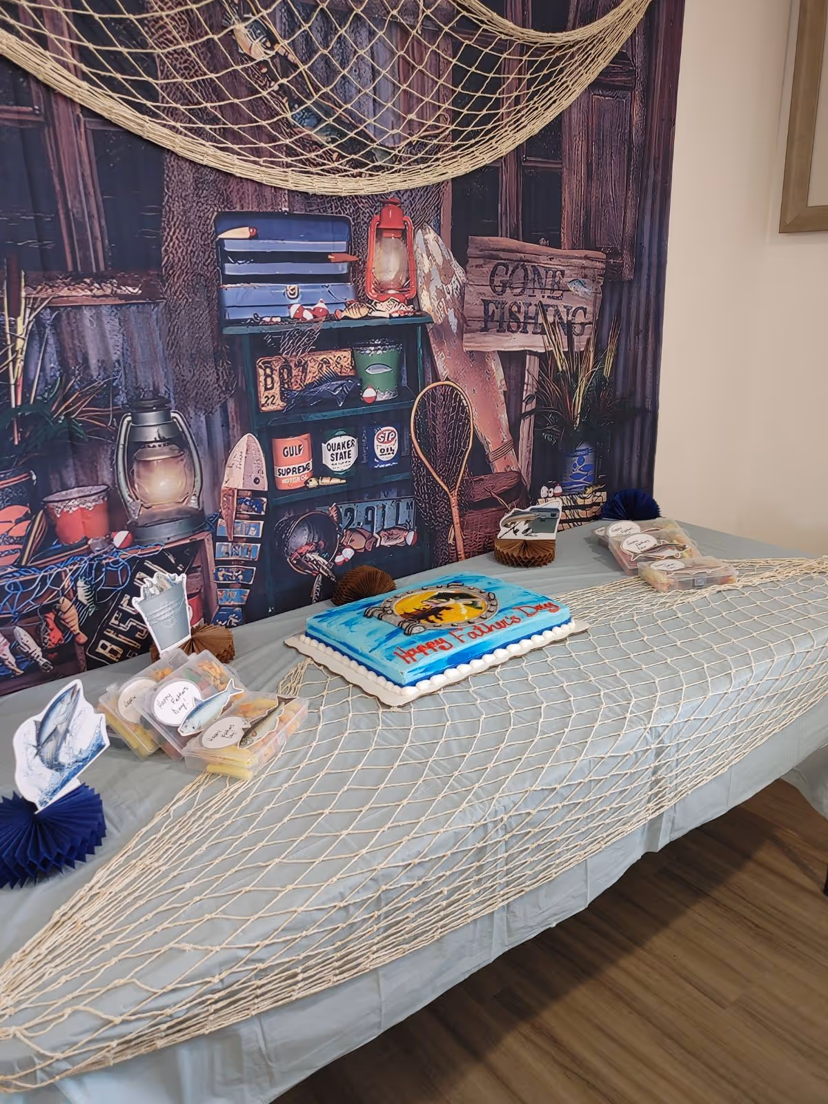 A fishing-themed indoor party table with a decorated cake, snacks, a fishing net on the table, and a backdrop that says 'Gone Fishing'.