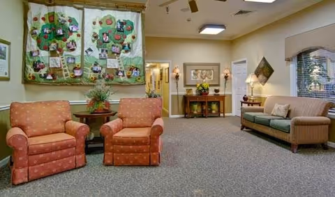 A cozy living room area in a senior living facility with two orange patterned armchairs and a small round table with a plant between them on the left. On the right, there is a beige sofa with cushions. The walls are decorated with a large quilted wall hanging featuring photos and notes, a framed picture, and a small table with flowers and lamps. The room has carpeted flooring and soft lighting.