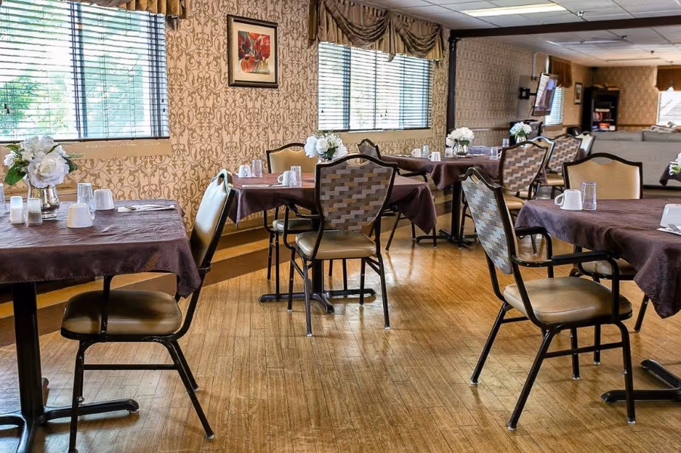 A dining room with several tables covered in brown tablecloths, each set with white cups, glasses, and small flower arrangements. The room has large windows with blinds, patterned wallpaper, and wooden flooring. Chairs with cushioned seats and patterned backs are arranged around the tables.