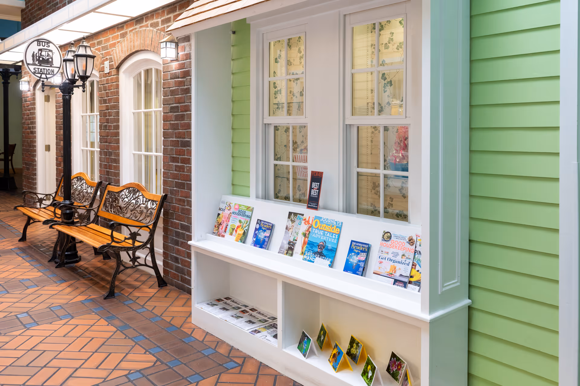 Indoor corridor styled like a small-town street with benches, a bus station sign, windows and a magazine display against green siding.