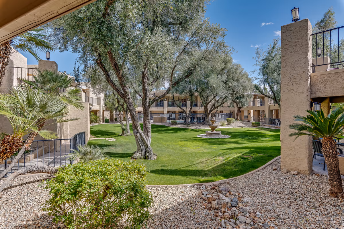 Outdoor courtyard area of Scottsdale Village Square featuring green grass, several trees, a central water fountain, and surrounding beige stucco buildings with balconies under a clear blue sky.