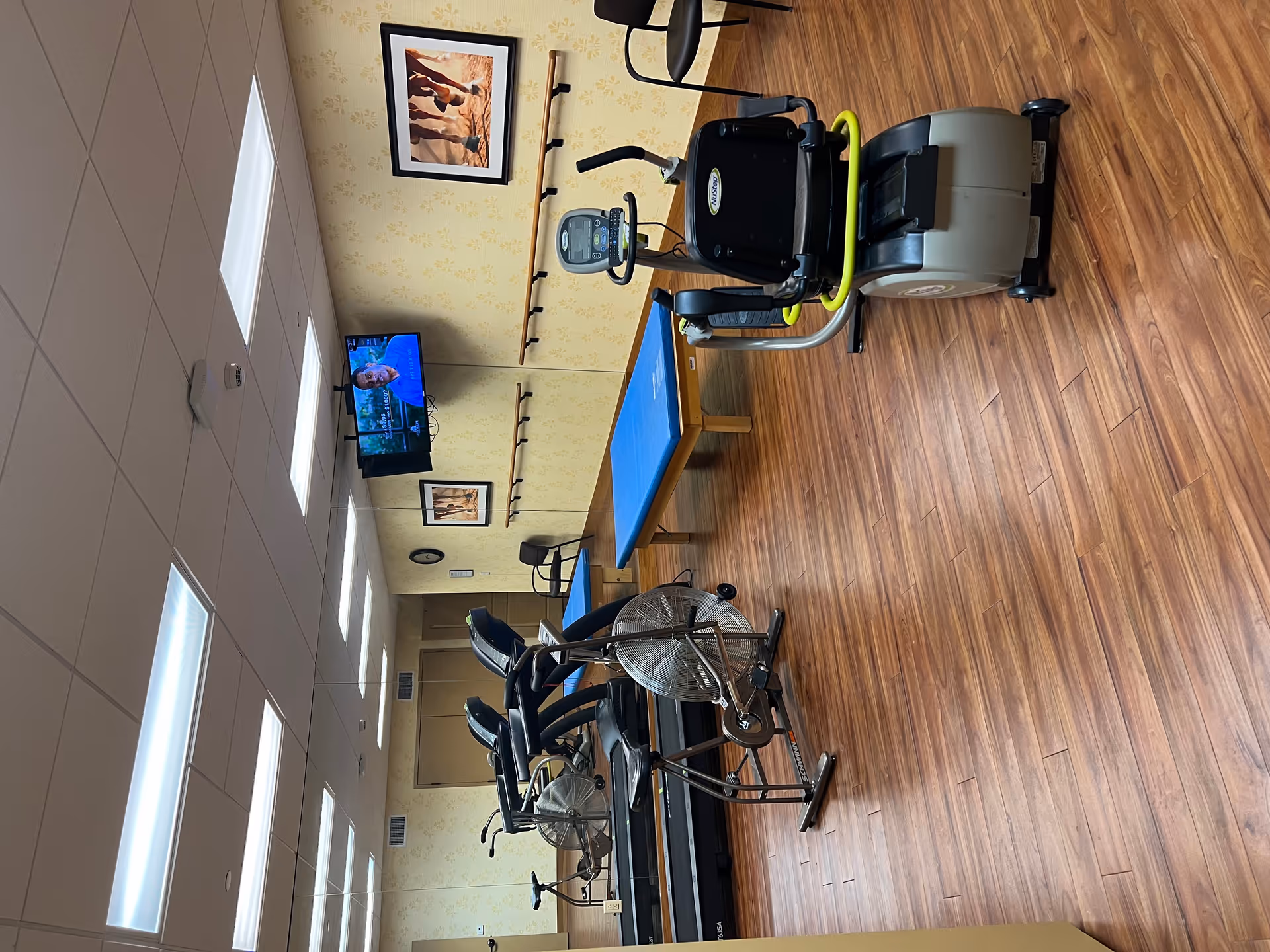 A fitness room with wooden flooring featuring exercise equipment including a recumbent bike, two treadmills, and a stationary bike. There is a blue padded exercise table against the wall, a wall-mounted TV showing a man speaking, two chairs, a clock, and framed pictures of horses on the walls. The room has a mirrored wall and fluorescent ceiling lights.