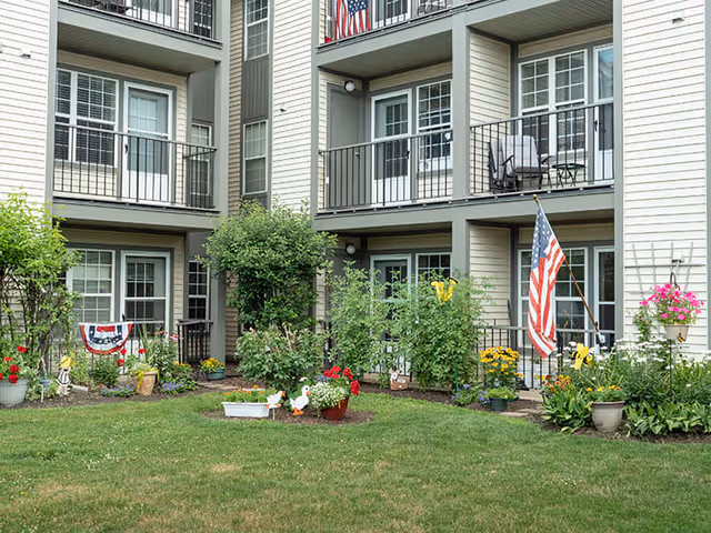 Exterior view of a multi-story residential building with balconies and patios. The ground level features a garden area with various plants, flowers, and decorative items including an American flag and garden ornaments. The building has beige siding and white-framed windows.