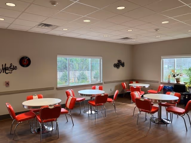 A bright dining room with several round white tables and red cushioned chairs. The room has large windows letting in natural light, wood flooring, and simple wall decorations including a clock and a wall art piece spelling 'Music'.