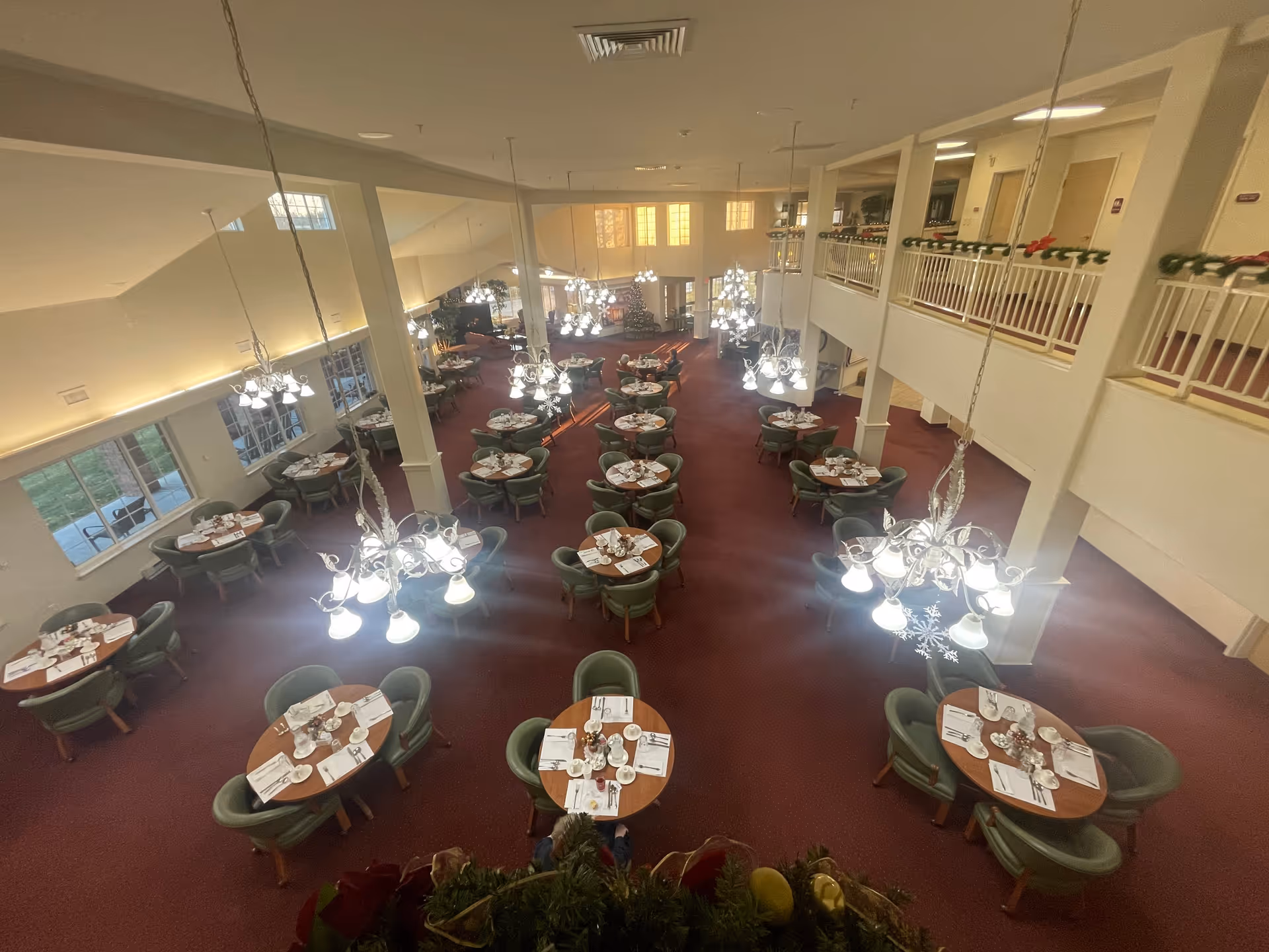A large dining room with multiple round tables set with white napkins, plates, cups, and silverware. Each table is surrounded by green cushioned chairs. The room has red carpet flooring, white walls, and several chandeliers hanging from the ceiling providing bright lighting. There is a second-floor balcony with white railings decorated with garlands, and large windows letting in natural light.