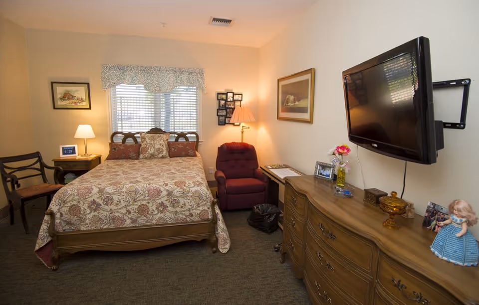 A cozy bedroom with a wooden bed covered in a floral patterned bedspread, a wooden dresser with a mounted flat-screen TV above it, a burgundy armchair with a floor lamp beside it, a small side table with a lamp and digital clock, and framed pictures on the walls. A window with blinds and a decorative valance is behind the bed.