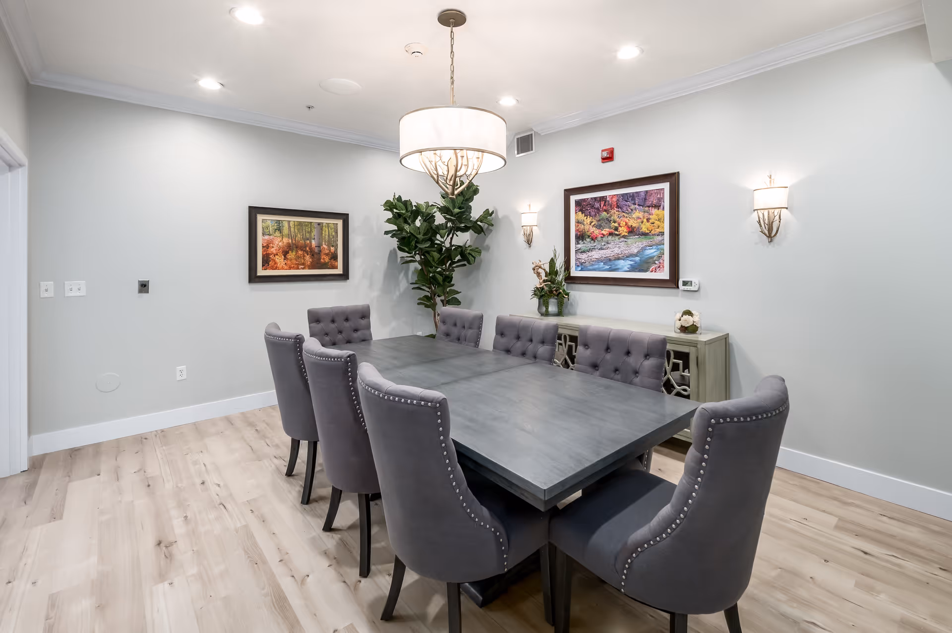 A modern dining room with a large rectangular dark wood table surrounded by eight gray upholstered chairs with tufted backs and nailhead trim. The room has light wood flooring, light gray walls, two framed landscape paintings, a large green plant in the corner, a sideboard with decorative items, and a round chandelier hanging above the table.