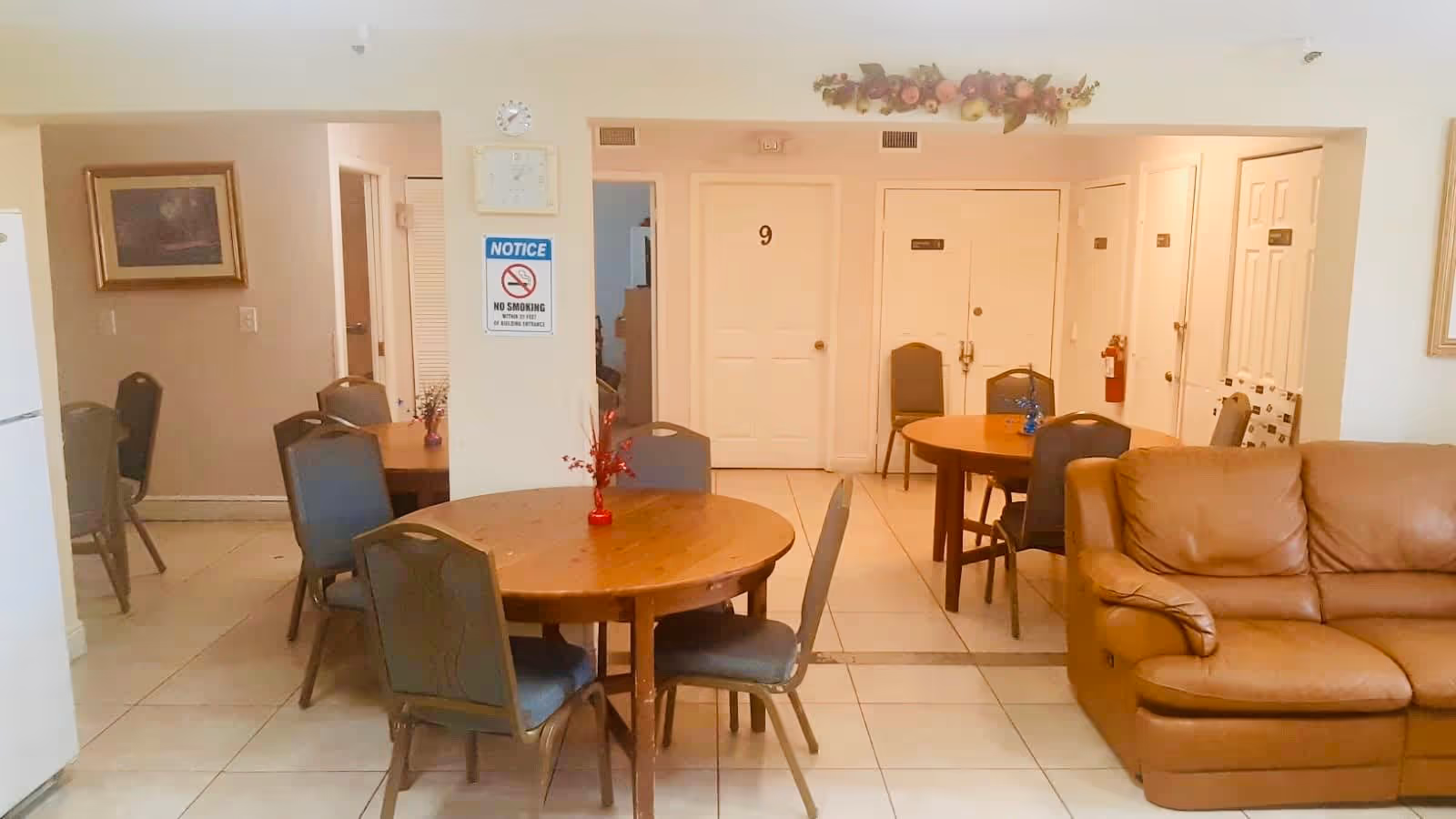 Community dining area with round wooden tables and chairs, a tan leather sofa, and numbered doors in the background.