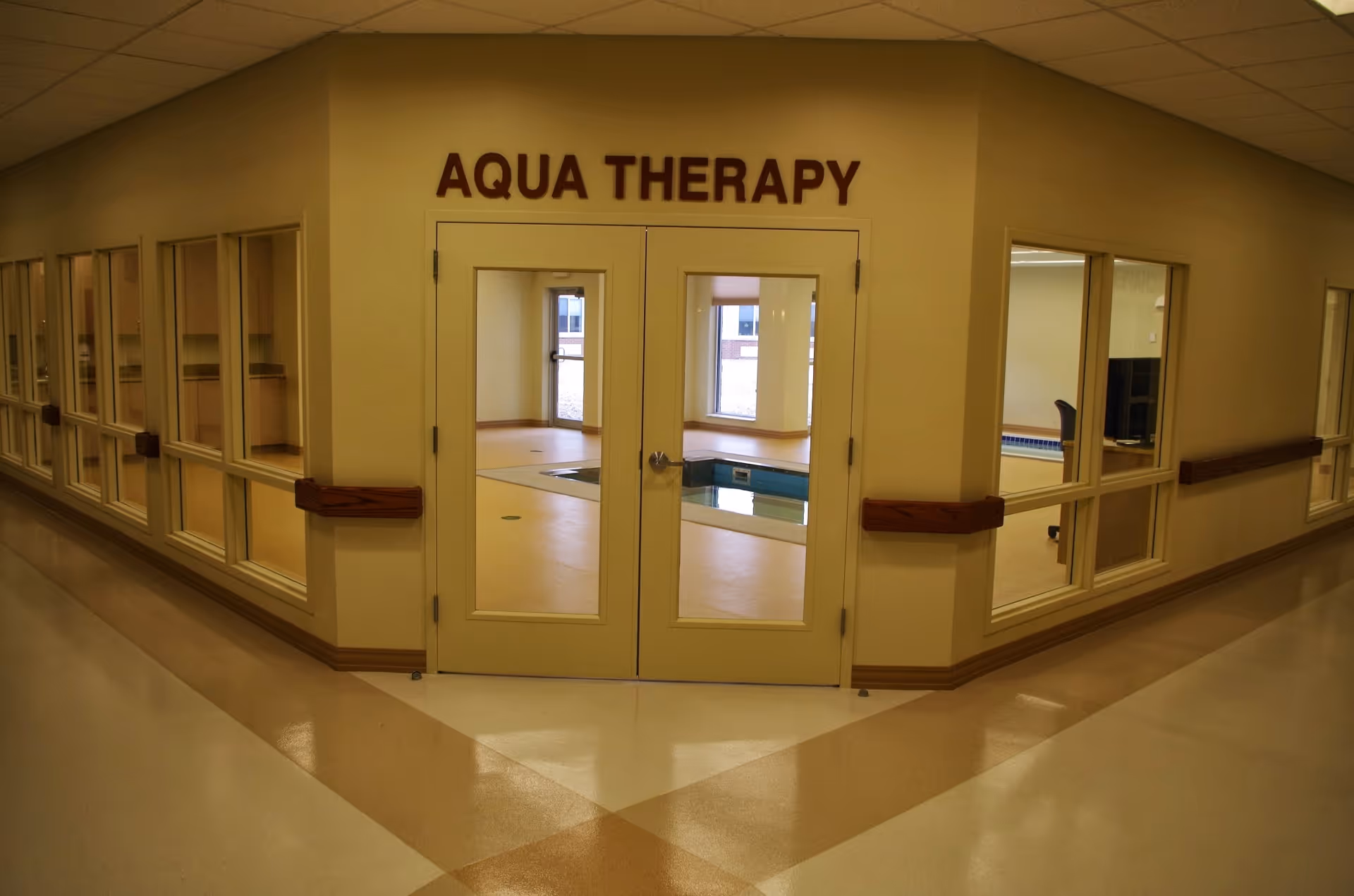 Double doors labeled "AQUA THERAPY" opening to an indoor therapy pool visible through windows in a hallway.