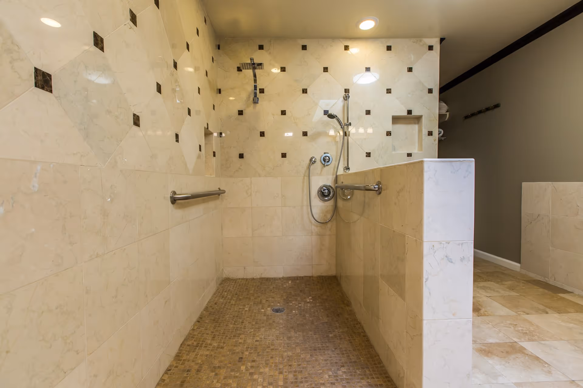 Spacious tiled roll-in shower with grab bars, handheld and overhead showerheads and a half-height partition.