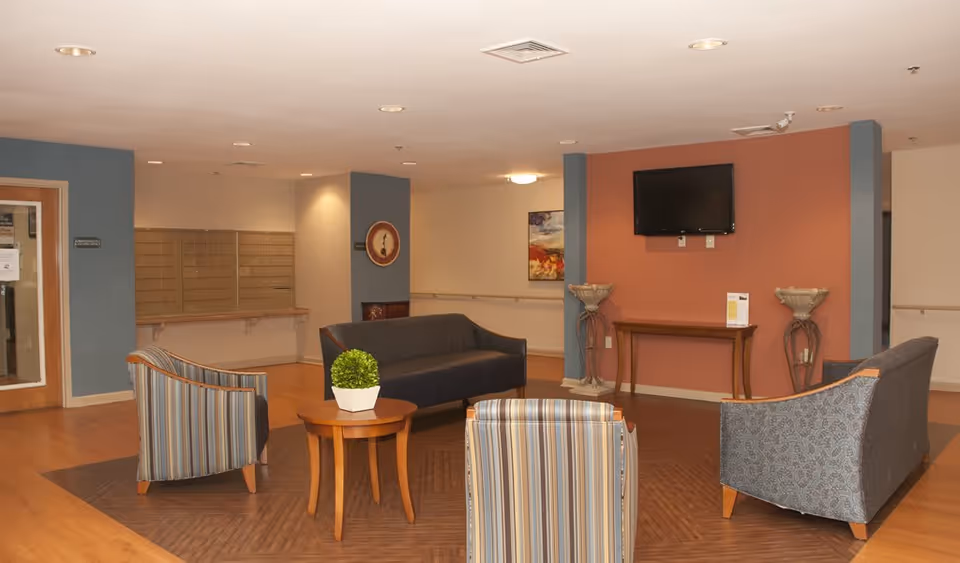A seating area in a senior living facility with four upholstered chairs arranged around a small wooden table with a potted plant. The room has wood flooring, a wall-mounted TV, decorative wall art, and a set of mailboxes along one wall.