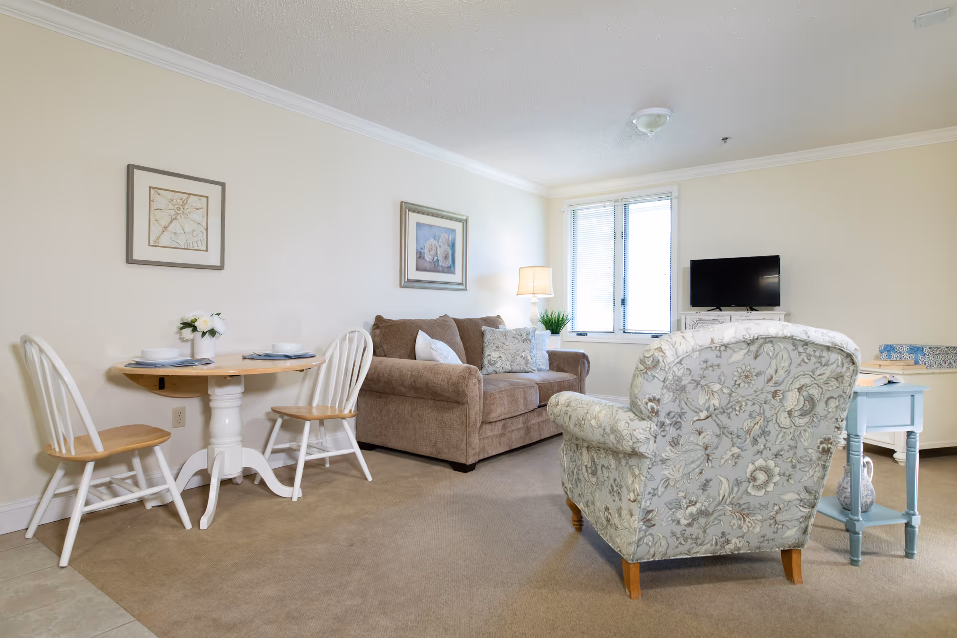 Bright, cozy living room with a sofa, floral armchair, small round dining table with chairs, TV on a stand, and a window.