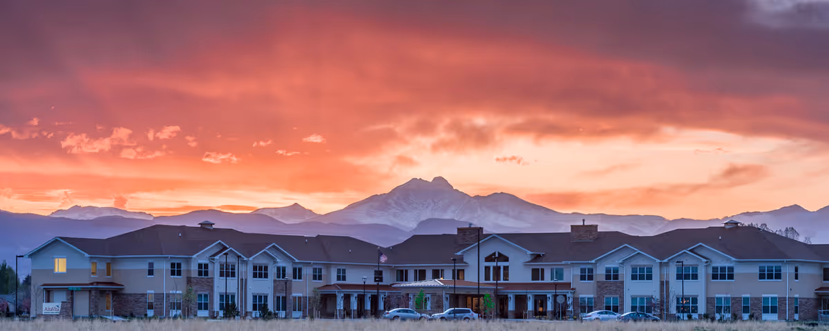 Front exterior of an assisted living building with parked cars, set against mountains beneath a dramatic orange and purple sunset.